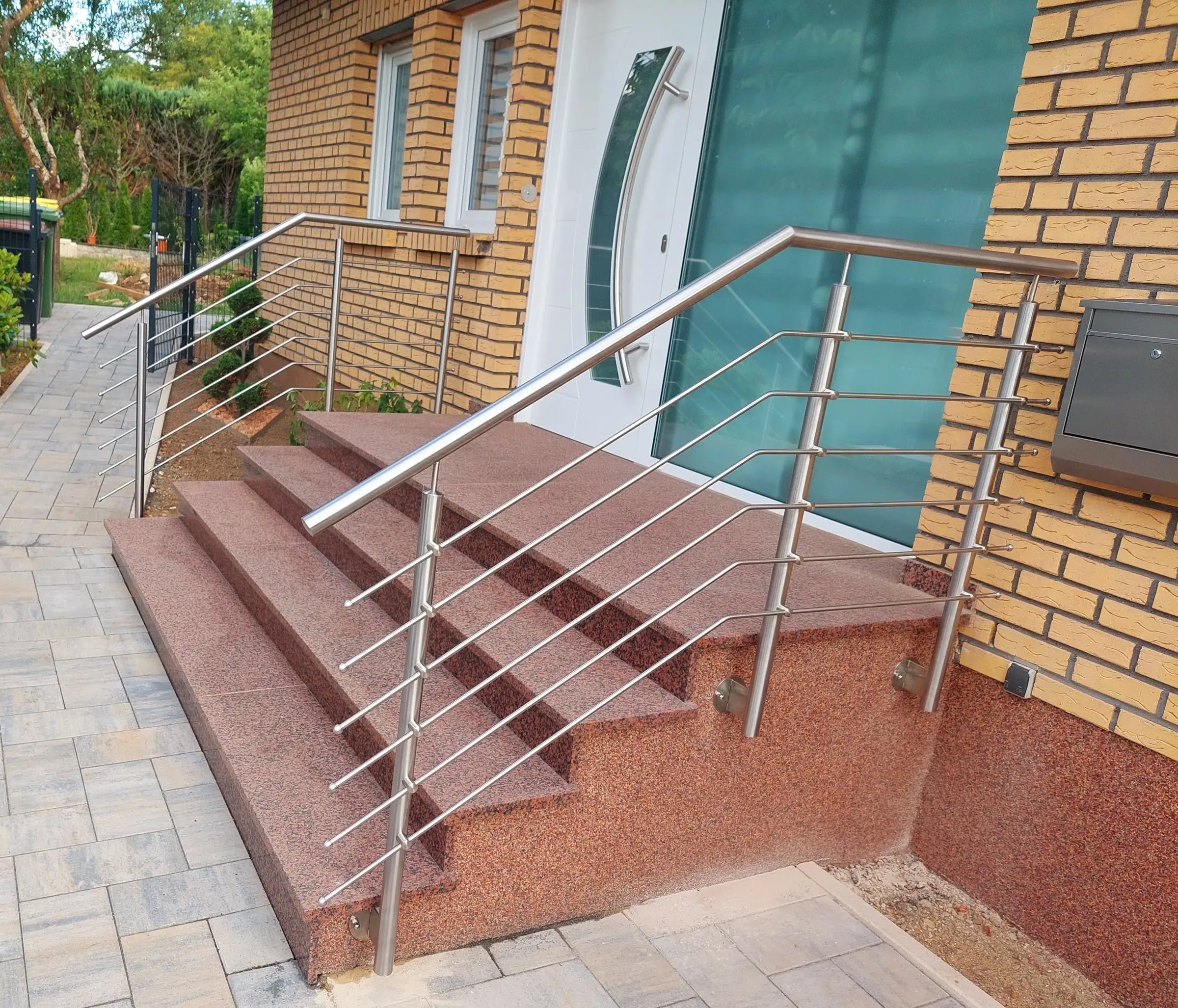 Zwei Edelstahlgeländer mit waagerechten Stäben an einer roten Granittreppe vor einer Haustür