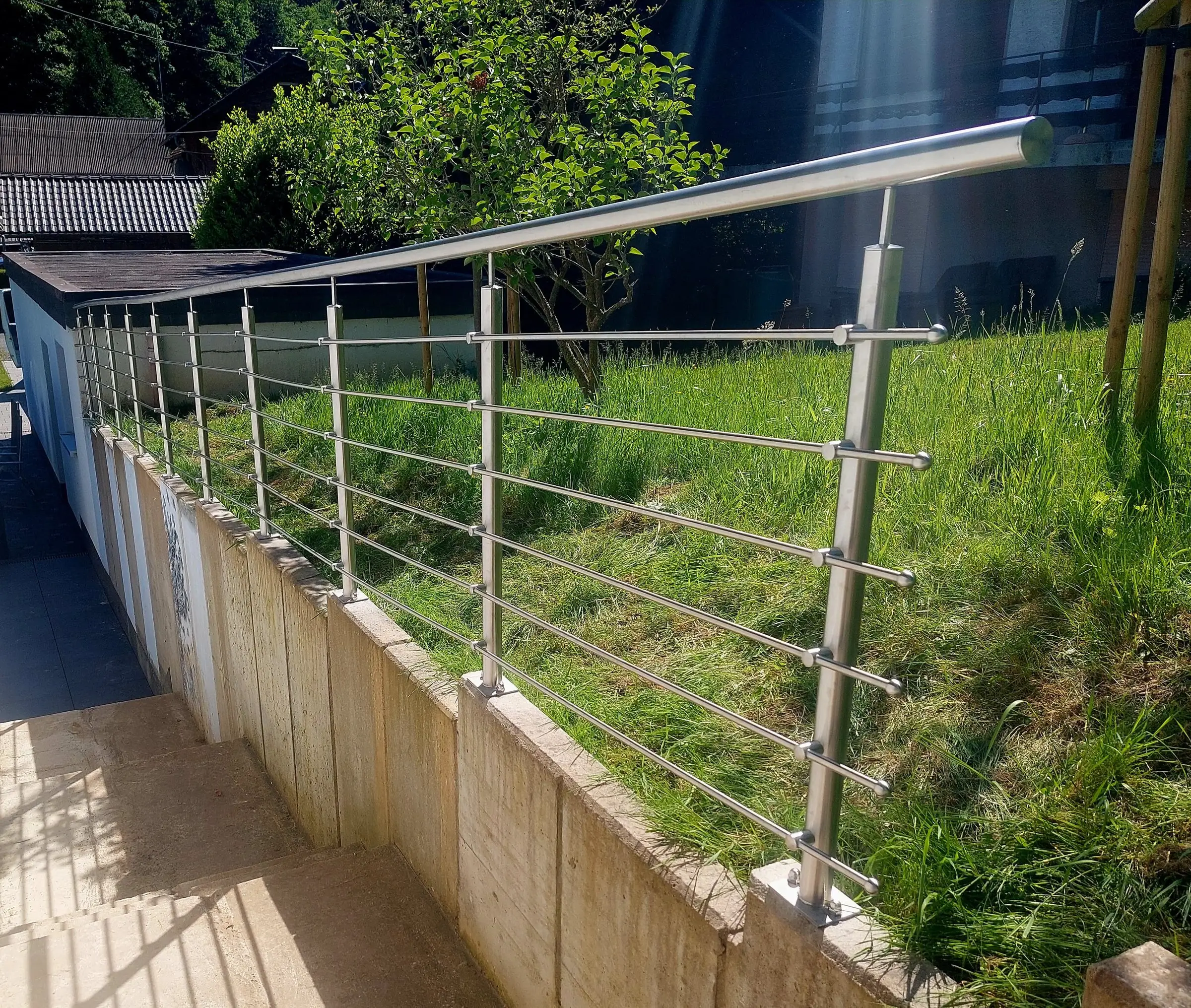 Langes Edelstahlgeländer mit waagerechten Stäben auf einer Betonmauer neben einer Außentreppe, dahinter Rasen